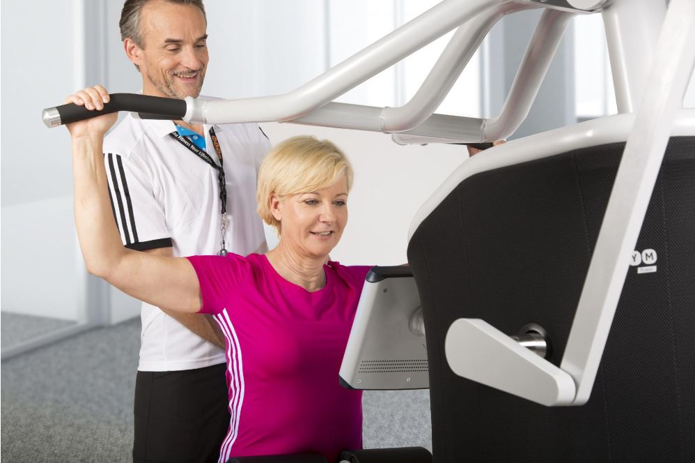 Oudere blonde vrouw met roze shirt krijgt instructie van eGym coach op eGym toestel.
